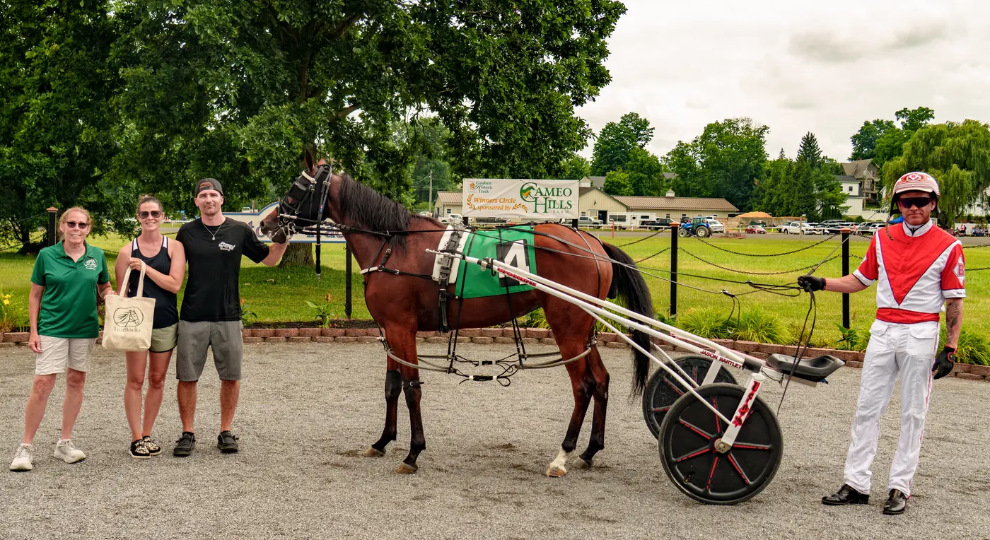 The winner's circle at Goshen Historic Track in Goshen, New York, after the Landmark Stake for two-year-old fillies on July 5th 2026. MissNeverDoNoWrong stands in harness with her connections and with driver Jason Bartlett in red silks.